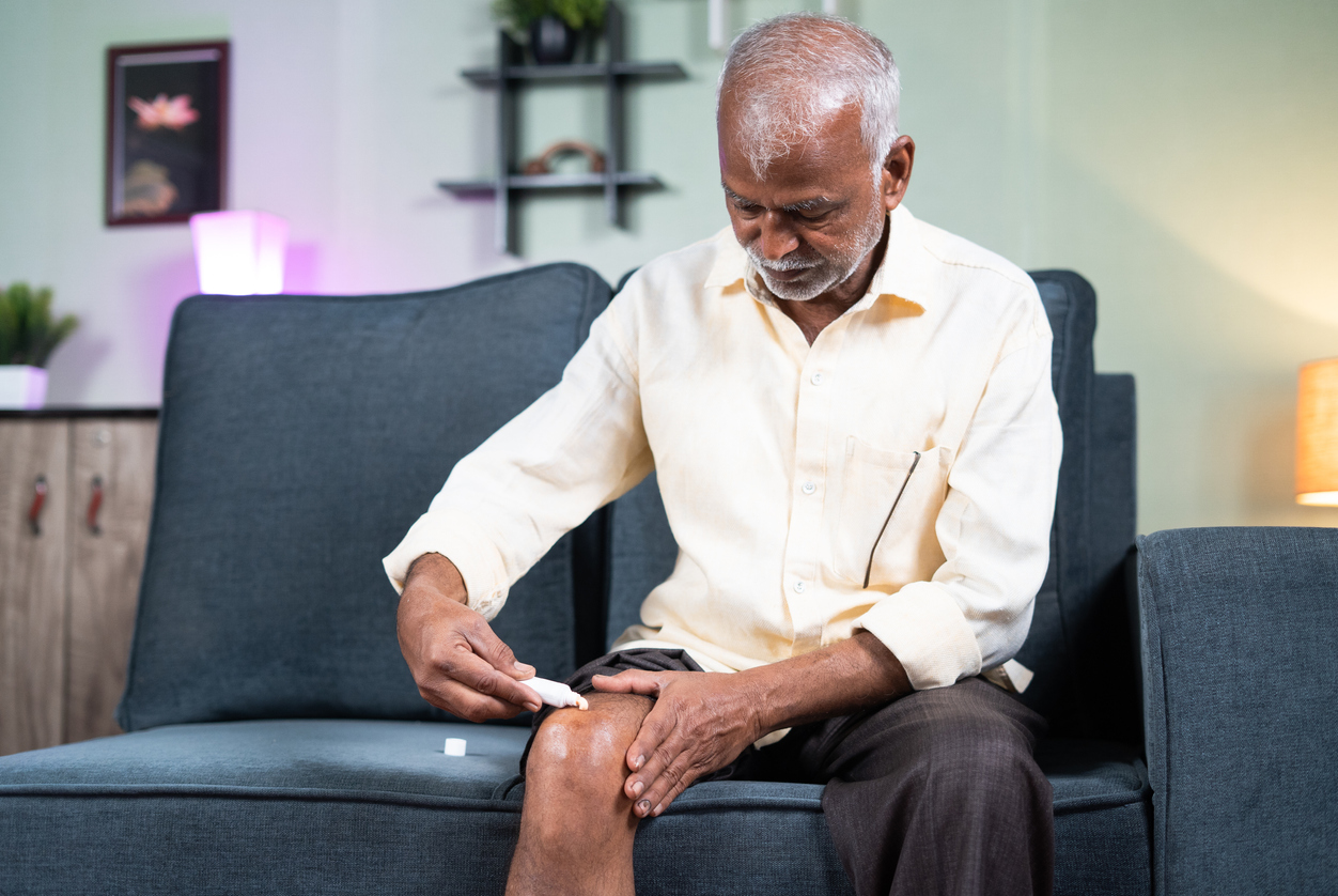 Focus on hands, Senior man applying ointment cream for joint knee pain at home while sitting on sofa – concept of treatment for osteoarthritis, knee injury and arthritis