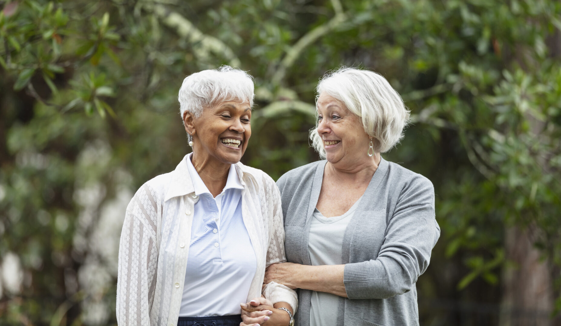 Two multiracial senior women engage in conversation while walking together outdoors in a backyard, with trees in the background.