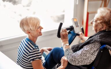 Therapist Helping Senior Woman On Wheelchair Doing Exercises On Exercising Bike