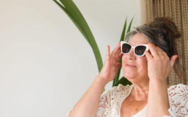 retired gray-haired senior adult woman putting on a pair of sunglasses at her beach apartment to enjoy a summer season day.