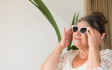 retired gray-haired senior adult woman putting on a pair of sunglasses at her beach apartment to enjoy a summer season day.