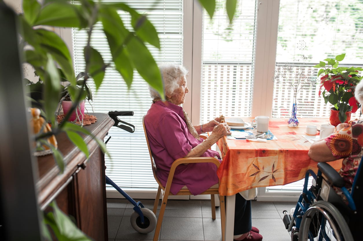 Elderly woman sitting at a table in a brightly lit room, featuring a walker and wheelchair nearby, illustrating considerations for dementia-friendly home design.