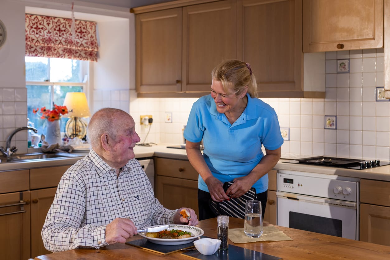 Caregiver smiling at an elderly man enjoying a meal, emphasizing the importance of proper nutrition in supporting cognitive health and fighting Alzheimer's disease.