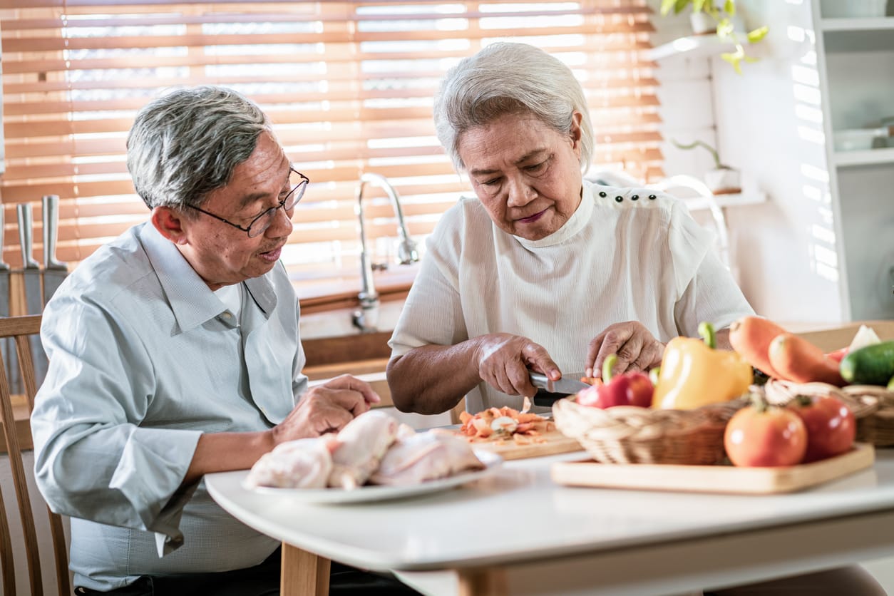 An elderly couple preparing a meal together, chopping vegetables in a bright kitchen, emphasizing the importance of incorporating protein into a balanced diet for seniors.