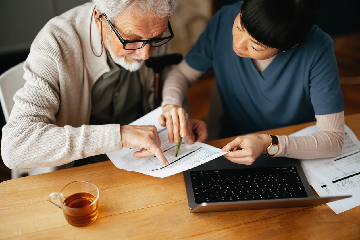 A senior man reviewing documents with a caregiver, highlighting the importance of understanding and accessing government benefits for senior care, with a laptop on the table for additional research.