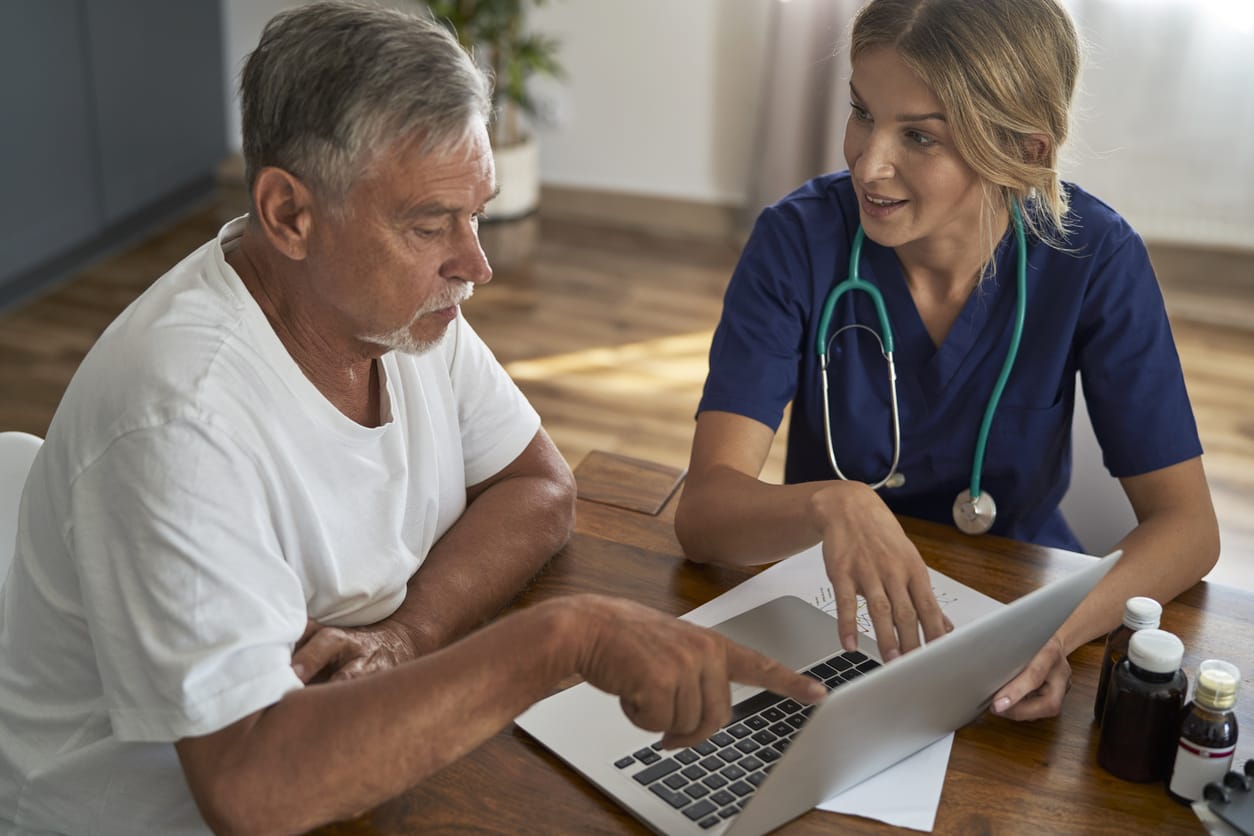 An older man with short grey hair is looking at a laptop with a doctor with long blond hair, blue scrubs and a stethescope.
