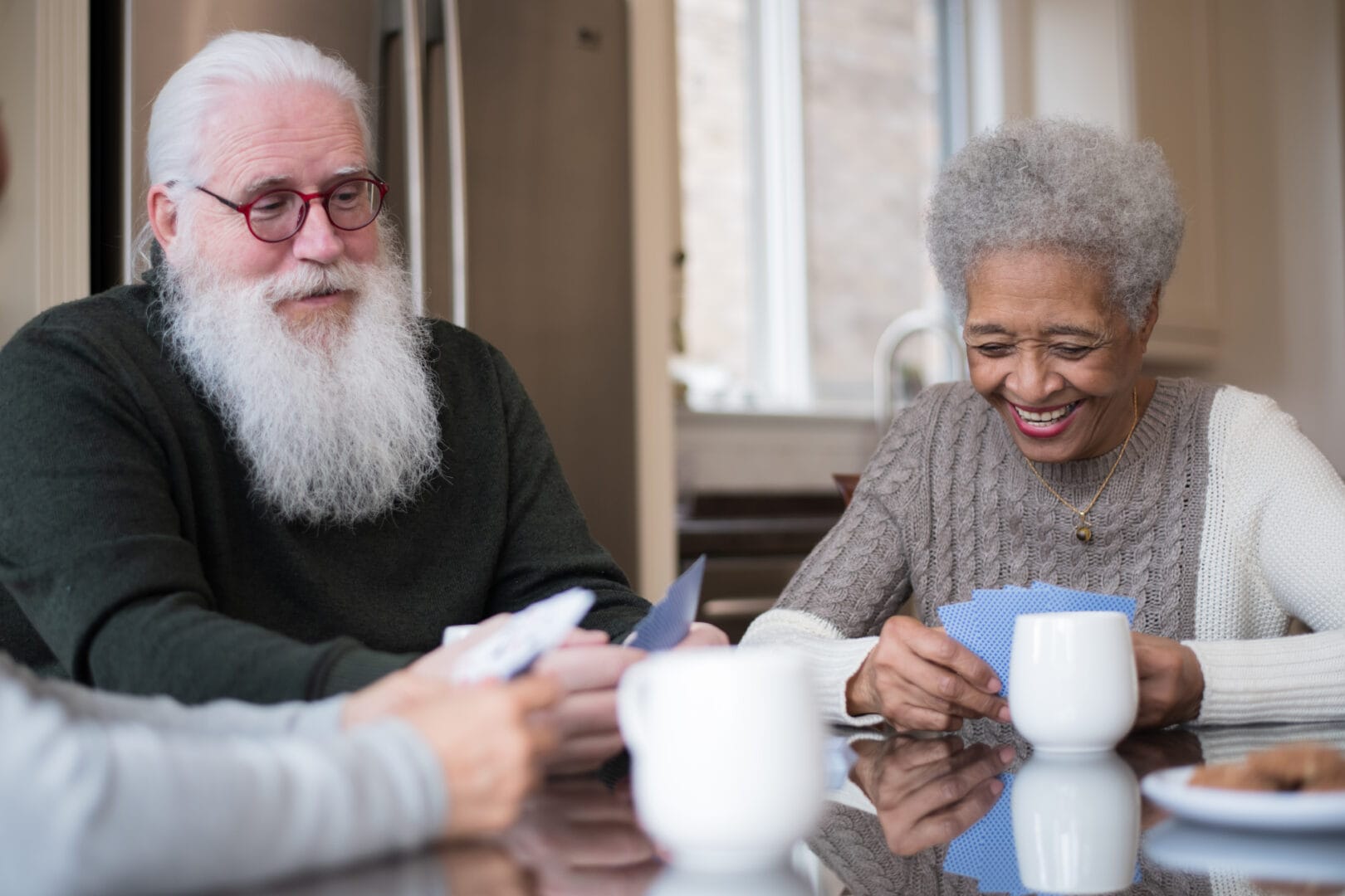 A senior Caucasian male with white hair and a long white beard, a senior woman with a beautiful smile wearing a sweater, and one other person who you can just see their arms and hands are sitting at a kitchen table drinking coffee and playing a card game together.