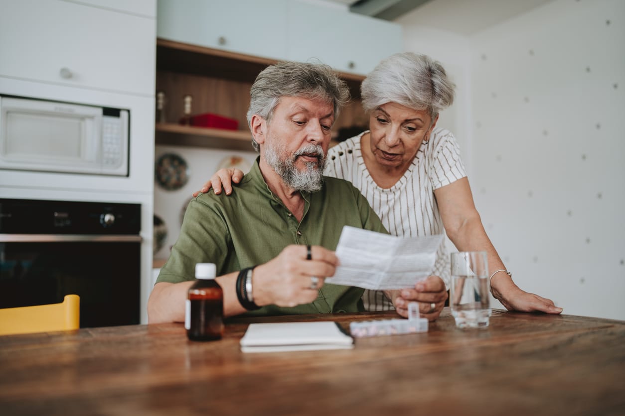Senior couple reading over financial documents at home in the kitchen with medications and a glass of water on the table.
