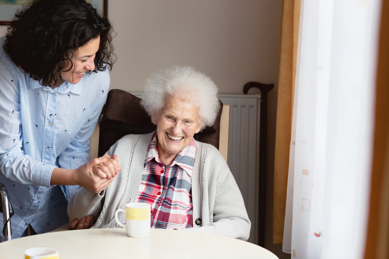 Celebrate National Centenarian's Day: Honoring 100 Years of Life. A young Hispanic woman is taking care for a senior woman, who is 94 years old and suffering from Dementia, the senior woman is wearing a checkered blouse and grey woolen jacket, she is sitting in an armchair in nursing home, she is laughing, holding the hand oft he caregiver.