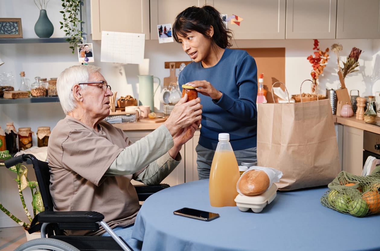 Safe Food Preservation & Storage for Seniors Volunteer delivering food for senior man who using wheelchair, they sitting in the kitchen and discussing food.