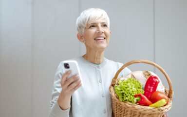 Beautiful senior woman holding basket full of fresh vegetables and using smart phone