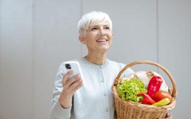 Beautiful senior woman holding basket full of fresh vegetables and using smart phone