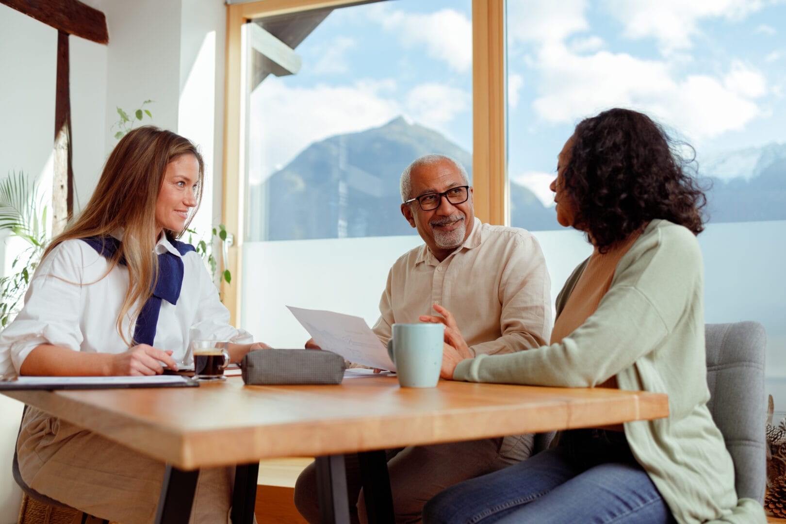 A financial advisor is seated with a senior couple in a bright room with a stunning mountain view. They are engaged in conversation about financial planning for long-term care, with documents laid out on the table.